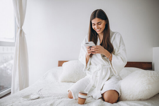 Young woman in bed in bathrobe with phone drinking coffee