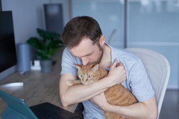 Caucasian male freelancer in gray t-shirt with cat. Portrait of cheerful bearded IT engineer at working place in home office sitting at table with tablet computer holding and kissing tabby cat