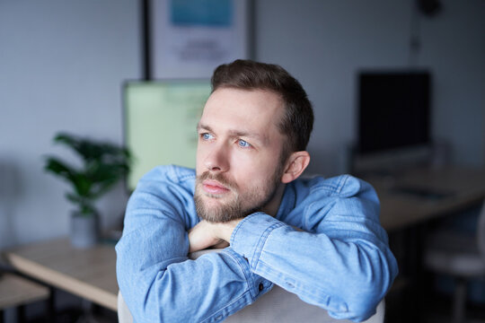 Cheerful bearded caucasian male sitting in home office looking at side dreaming. Young businessman or entrepreneur in denim shirt with computer monitor on background. High quality image