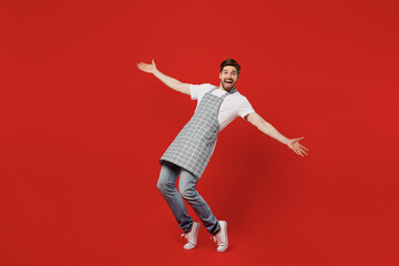 Full body side view happy young housekeeper chef cook baker man in grey apron look camera with outstretched hands stand on toes lean back isolated on plain red background studio Cooking food concept