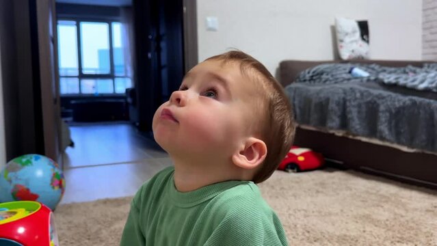 Little kid sits on the floor near his toys. Baby boy looks up with interest taking a finger into mouth. Close up side view.