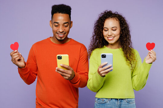 Young lovely couple two friends family man woman of African American ethnicity wear casual clothes together use mobile cell phone hold paper red heart isolated on pastel plain light purple background.