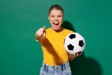 Young woman fan wear basic yellow t-shirt cheer up support football sport team hold in hand soccer ball watch tv live stream point finger camera on you scream isolated on dark green background studio.