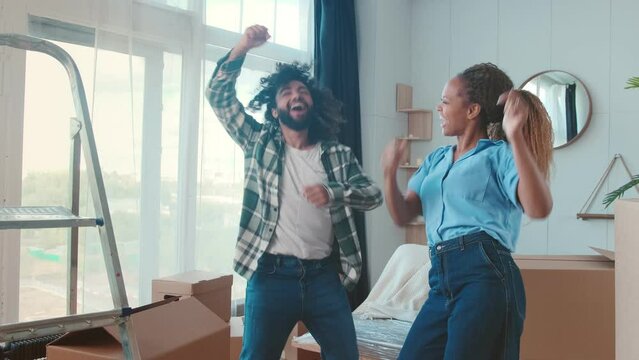 Young happy friends Arabian man and African American woman hosted house party in honor of successful purchase of cool property or start of renovation and dance in apartment standing among boxes