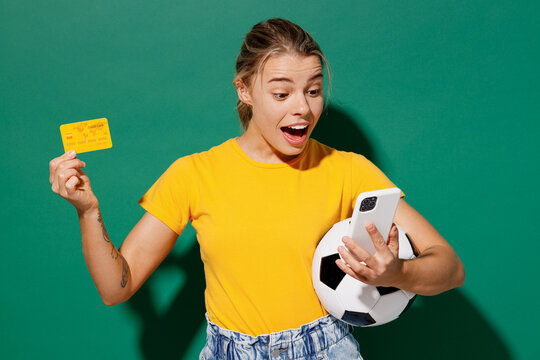 Young surprised woman fan wear basic yellow t-shirt cheer up support football sport team hold soccer ball mobile cell phone yellow card watch tv live stream isolated on dark green background studio.