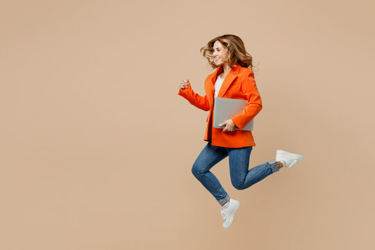 Full body side view young employee business IT woman corporate lawyer in classic formal orange suit glasses work in office jump high hold closed laptop pc computer isolated on plain beige background