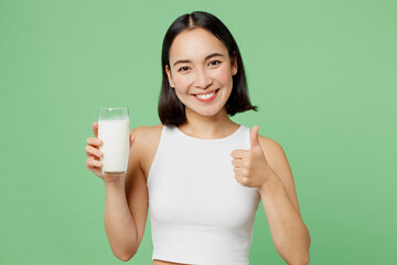 Young smiling happy woman wear white clothes hold in hand glass drink milk show thumb up isolated on plain pastel light green background. Proper nutrition healthy fast food unhealthy choice concept.