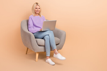 Full length photo of smart woman wear knit sweater jeans sit on armchair writing email remote job isolated on beige color background