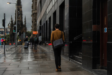 Young man in a brown sweater, walking down the city street with a black bag on a rainy day. 