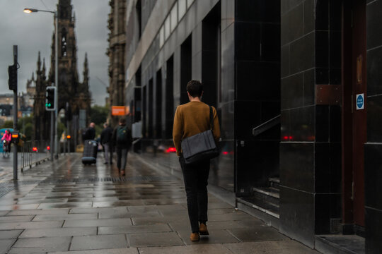 Young Man In A Brown Sweater, Walking Down The City Street With A Black Bag On A Rainy Day. 