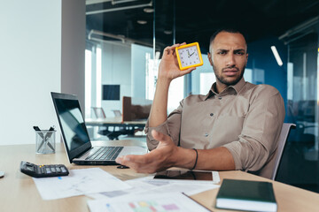 Dissatisfied hispanic man looking at camera and showing clock, businessman frustrated waiting for colleague to be late, working inside office with documents using laptop inside office. - obrazy, fototapety, plakaty