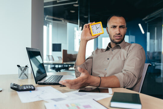 Dissatisfied hispanic man looking at camera and showing clock, businessman frustrated waiting for colleague to be late, working inside office with documents using laptop inside office.