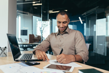 Fototapeta na wymiar Successful african american businessman inside office doing paperwork, man writing information, worker using laptop for work sitting with bills and contracts smiling and satisfied with result.