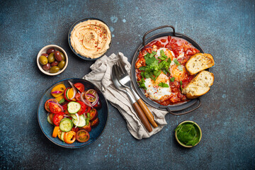 Fototapeta na wymiar Middle Eastern traditional breakfast or brunch with eggs Shakshouka in pan with toasts, fresh vegetables salad, hummus and olives on rustic concrete background table from above