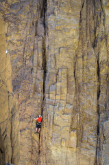 A male rock climber in a red t-shirt climbs a route on a steep granite wall on the rocks of the mountains