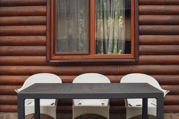 Three white rattan chairs and a table next to the wooden log house outdoors.