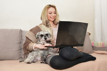 Lovely lady smiles and hugging a cute dog on the couch while watching movies on a laptop.