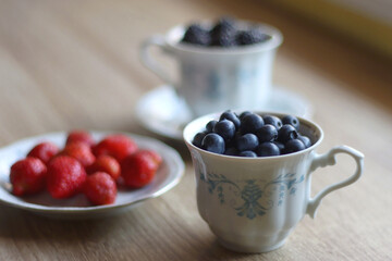 Vintage porcelain cups and plates filled with various fresh berries. Wooden background, selective focus.