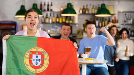 Group of emotional young adult fans supporting Portuguese sports team with state flag while resting in bar