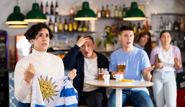 Emotional sad young female fan supporting team and holding state flag of Uruguay in hands during rest in sports bar
