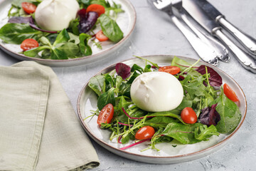 Plates with arugula and spinach salad with fresh cherry tomatoes and italian fresh cheese burrata with balsamic vinegar dressing on a light grey concrete background
