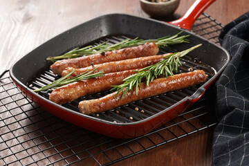 Traditional german organic pork sausages grilled on a red grill pan with rosemary branches and whole black pepper seeds on a baking rack on a wooden background