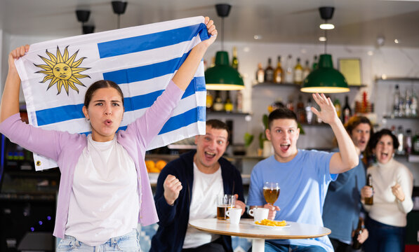 Screaming young adult sports fans rooting for favorite team and waving flag of Uruguay while watching match together in pub. High quality photo
