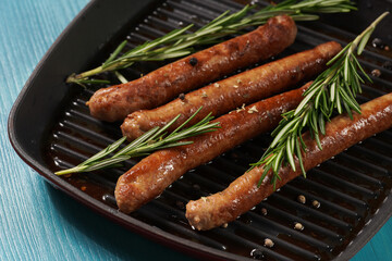 Traditional german organic pork sausages grilled on a red grill pan with rosemary branches and whole black pepper seeds on a baking rack on a wooden background, close up