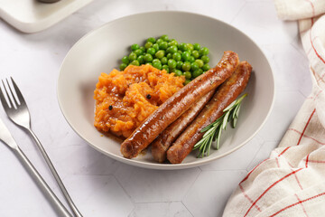 A lunch dish - boiled mashed sweet potato, steamed green peas and pork roasted sausages in a light grey bowl on a marble background
