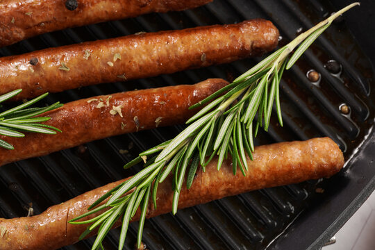 Traditional german organic pork sausages grilled on a red grill pan with rosemary branches and whole black pepper seeds on a marble background, close up