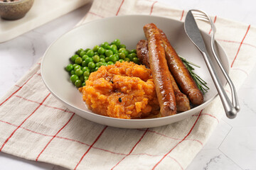 A lunch dish - boiled mashed sweet potato, steamed green peas and pork roasted sausages in a light grey bowl on a marble background