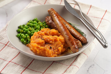 A lunch dish - boiled mashed sweet potato, steamed green peas and pork roasted sausages in a light grey bowl on a marble background