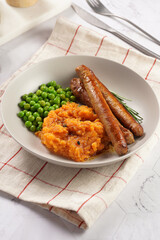 A lunch dish - boiled mashed sweet potato, steamed green peas and pork roasted sausages in a light grey bowl on a marble background
