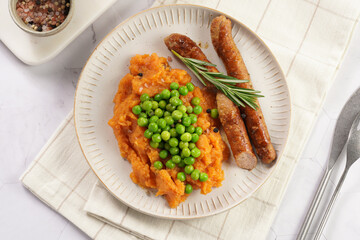 A lunch dish - boiled mashed sweet potato, steamed green peas and pork roasted sausages in a light grey bowl on a marble background, top view