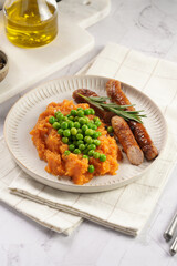 A lunch dish - boiled mashed sweet potato, steamed green peas and pork roasted sausages in a light grey bowl on a marble background