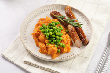 A lunch dish - boiled mashed sweet potato, steamed green peas and pork roasted sausages in a light grey bowl on a marble background