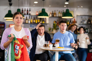 Fototapeta na wymiar Emotional sad young female fan supporting Portuguese team and holding state flag in hands during rest in sports bar