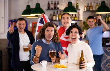 Obraz premium Happy young adult fans waving British flag while drinking beer and watching match together in sports bar