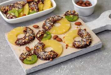 Chocolate covered pineapple rings and kiwifruit slices sprinkled with chopped nuts and coconut flakes on white wooden board. Homemade healthy fruit treat. Soft focus, horizontal.