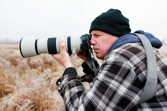 A photographer wearing winter clothing looks through the viewfinder of his camera to photograph his subject