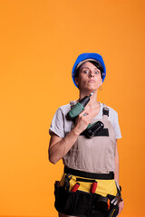 Female repair contractor holding power drill in front of camera, using electric drilling tool before renovation project. Construction worker wearing blue helmet and building uniform.
