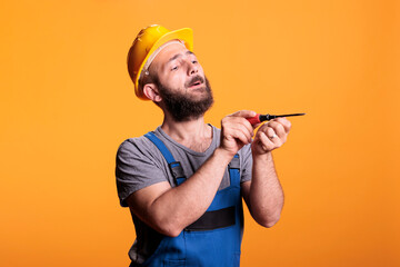 Building craftsman using screwdriver to drill nails, carpenter dressed in overalls with hardhat and tools belt. Professional repairman using tool on background in studio shot, industrial skill.