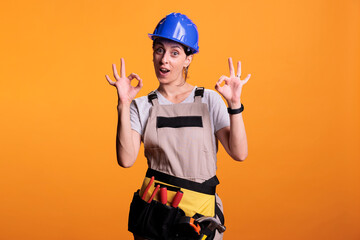 Young female contractor doing okay good symbol in studio, wearing renovating uniform and helmet. Cheerful construction worker showing approval and agreement gesture, achievement.