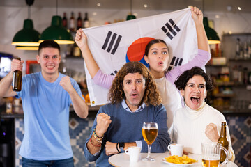 Company of young adult fans supporting team of South Korea with state flag while resting in sports...