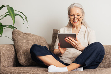 Smiling senior woman sitting with crossed legs using a digital tablet. Aged female in eyeglasses...