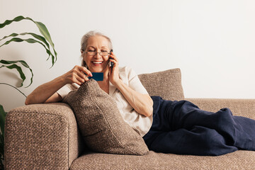 Happy senior woman holding a credit card and making call. Aged woman in glasses lying on sofa and...