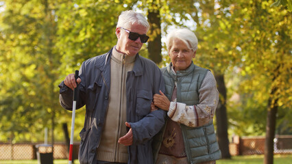 Senior woman helping her blind husband in sunglasses to walk through the park. High quality photo