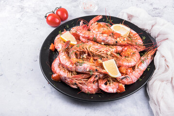 Baked langoustines in garlic sauce with cherry tomatoes on a black plate on a light marble background, side view. 