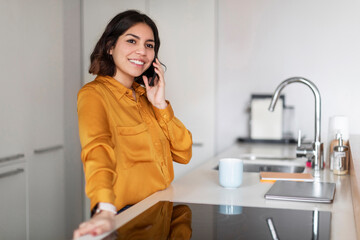 Portrait Of Smiling Young Arab Woman Talking On Cellphone In Kitchen Interior