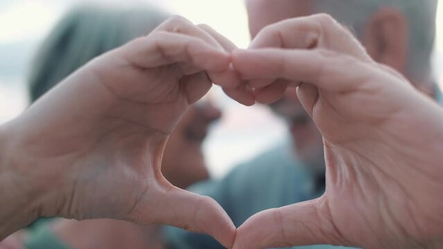 Close up portrait happy sincere middle aged elderly retired family couple making heart gesture with fingers, showing love or demonstrating sincere feelings together outdoors, looking at camera.
